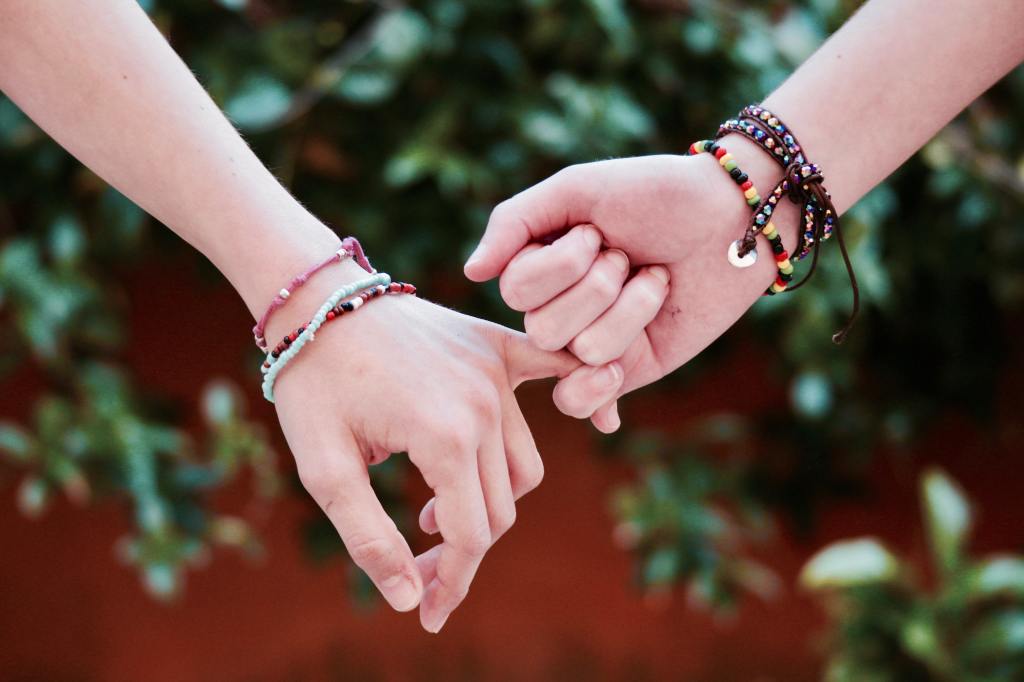 two female hands locking pinky fingers and wearing bead bracelets with a green tree background 