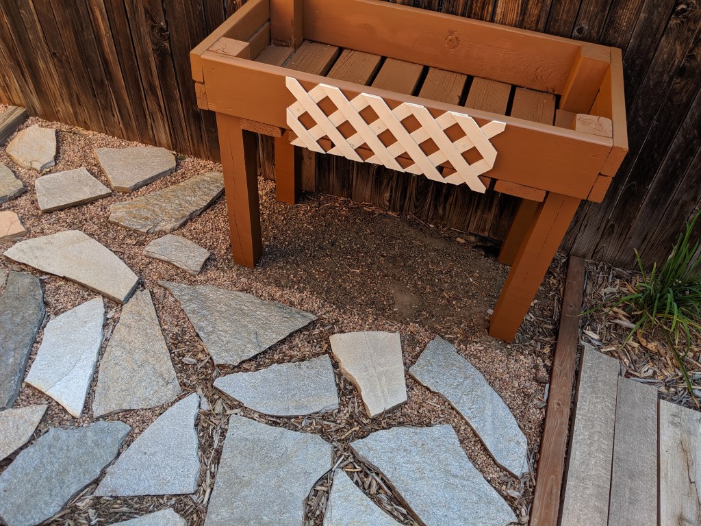 woodworking art by writer Vilma Reynoso, standup, rectangular, flower planter, brown, with white lattice decoration in front, set in a rock garden