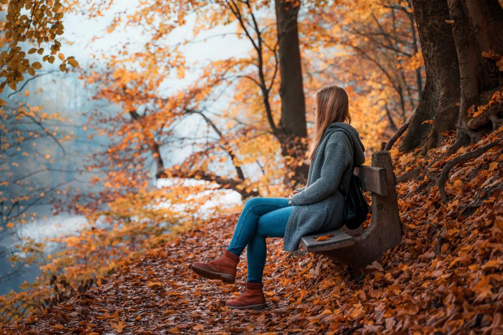 a girl sitting on a bench near trees in autumn with colorful leaves on the ground 