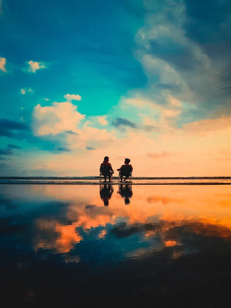 two people sitting on the shore on the beach side by side. One woman and one man on chairs 