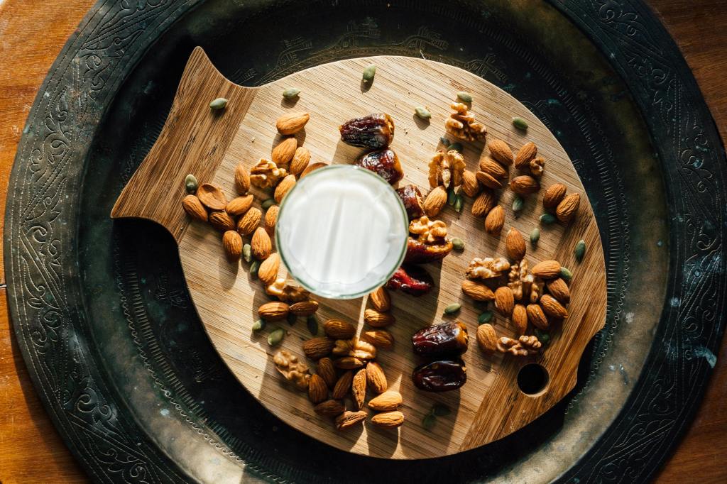 glass with nut milk in it over a circular wood platter with almonds and other nuts 